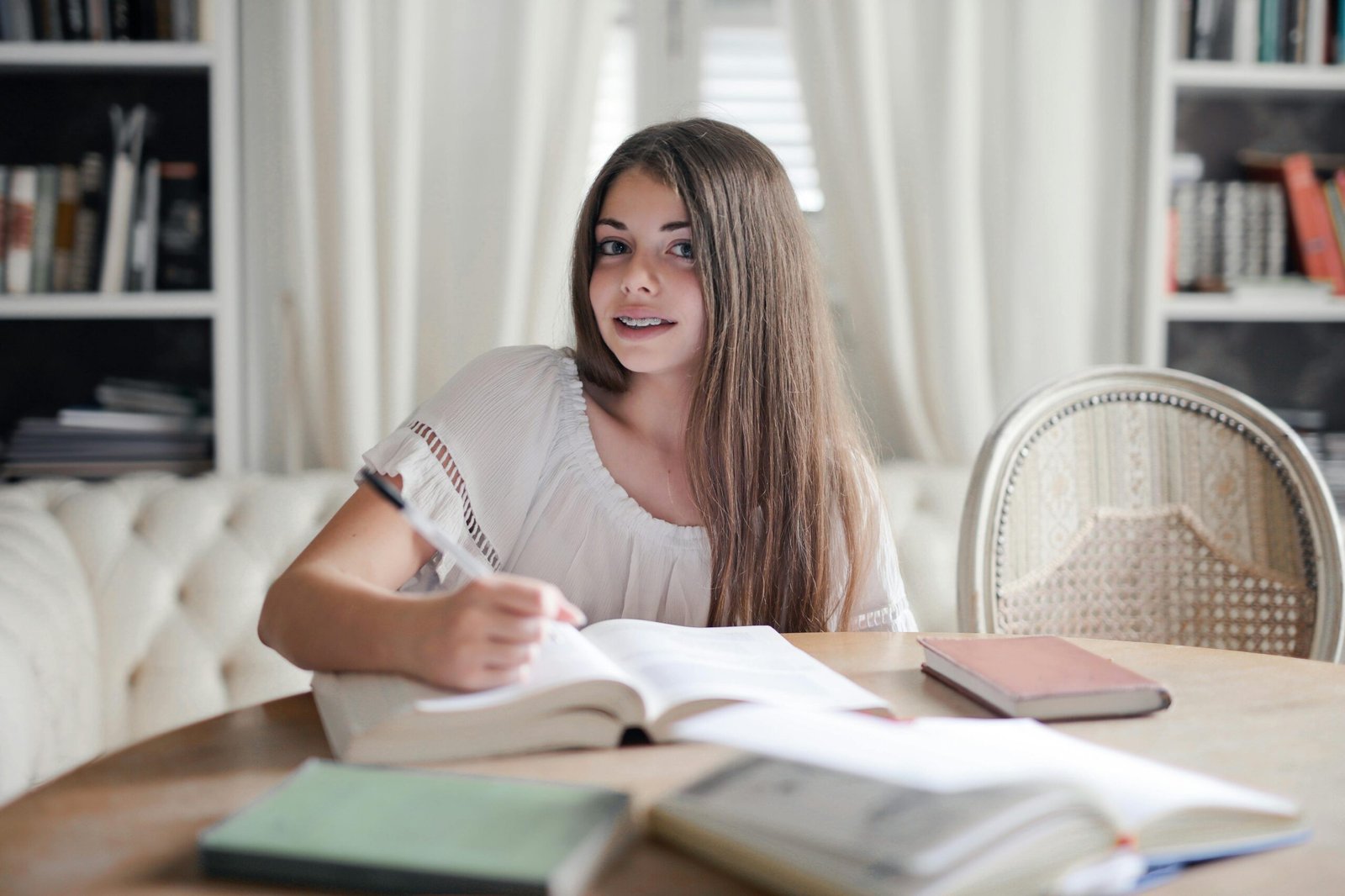 Smiling woman with long hair studying indoors at a table surrounded by books.