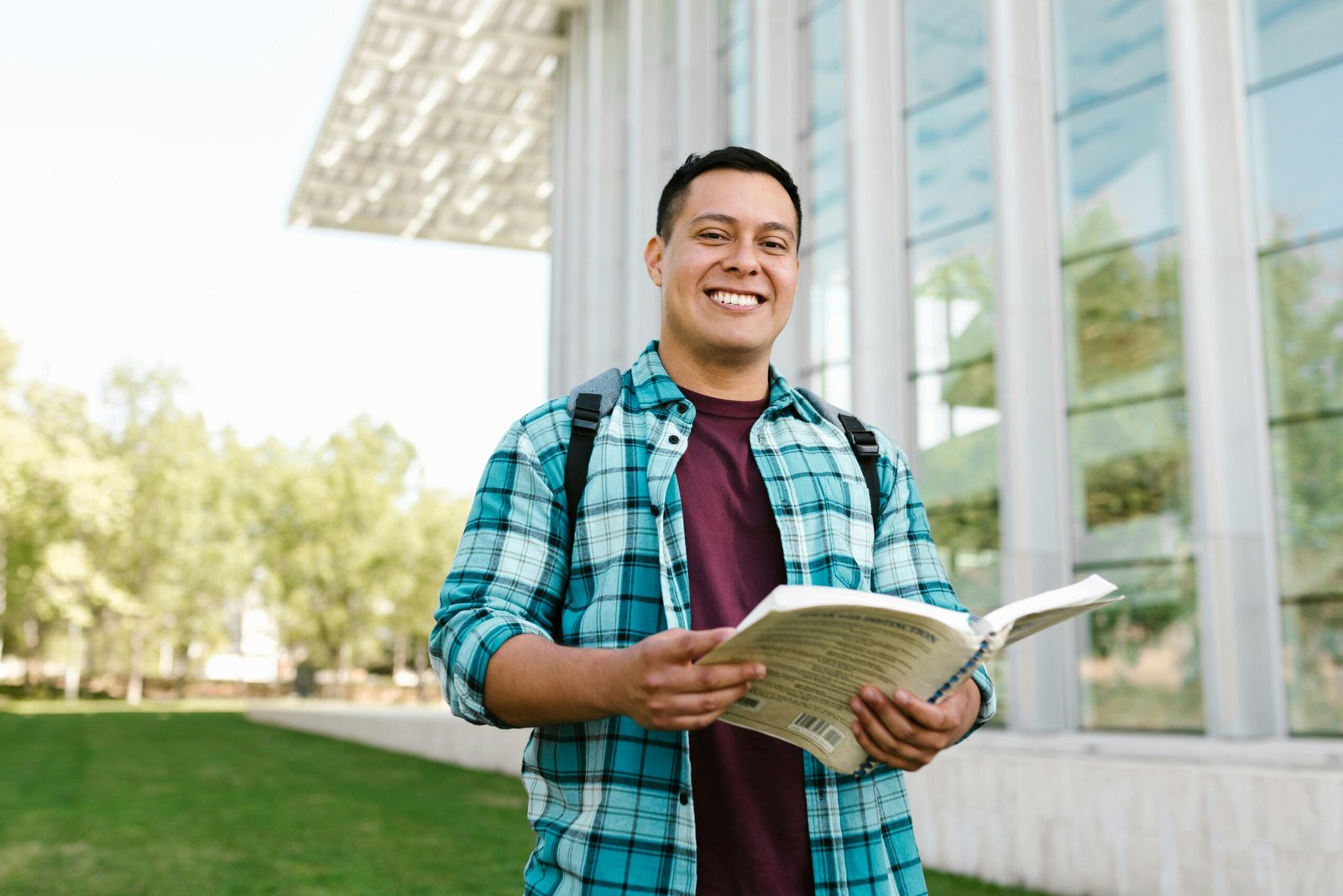 Young male student in plaid shirt smiling while holding a book outside a building.